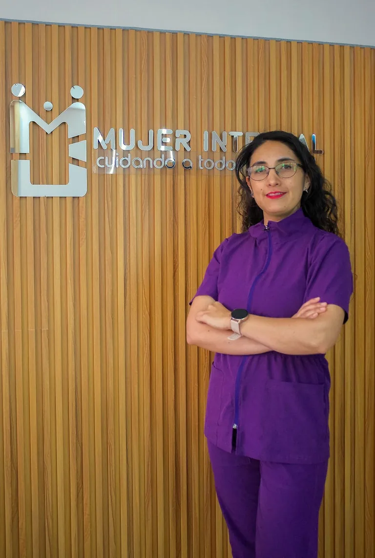 Bioquímica con uniforme morado, de pie con los brazos cruzados junto al rótulo Mujer Integral